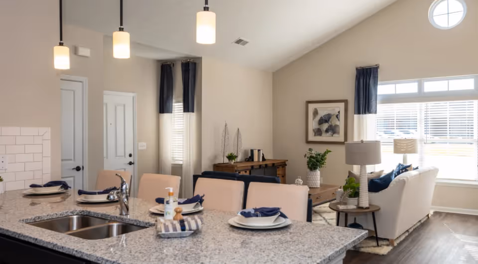 Open-plan interior showing a kitchen island with sink and place settings opening onto a bright living room with a sofa and large windows.
