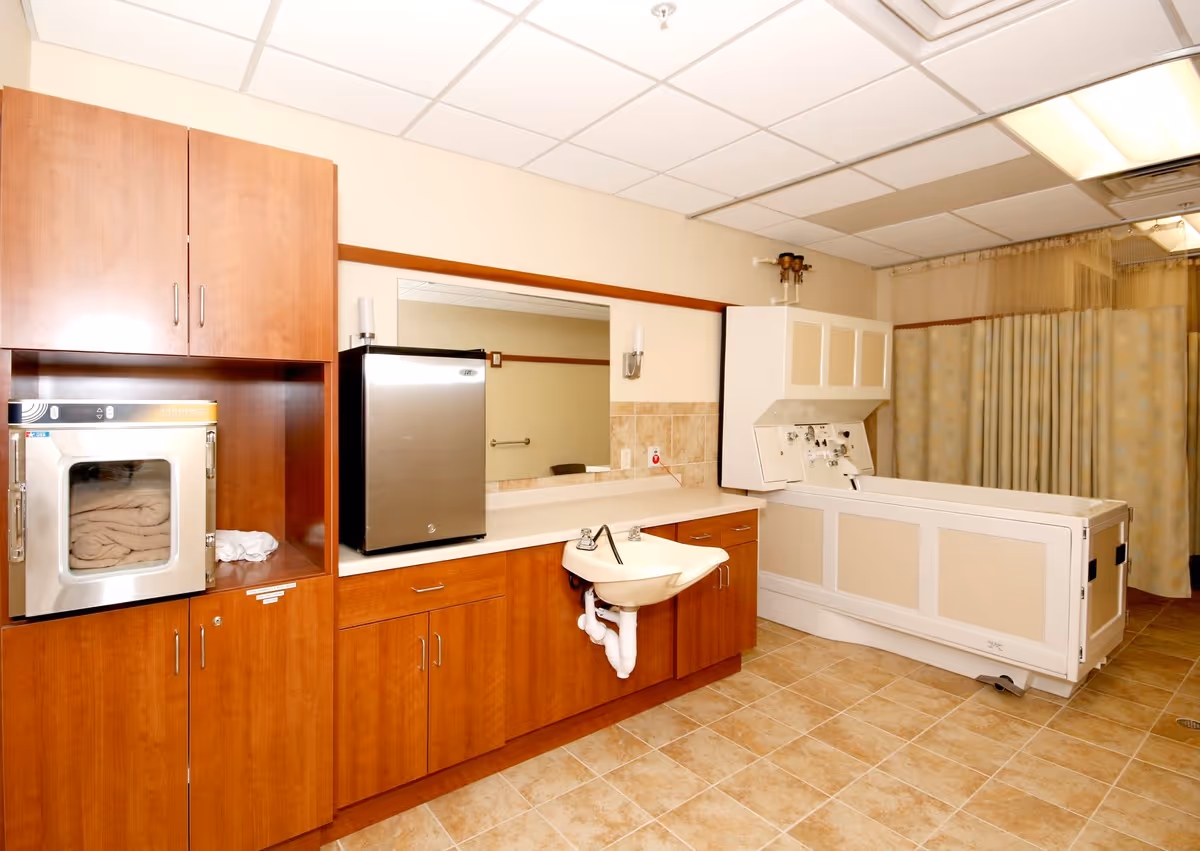A clean and well-lit room with wooden cabinets, a small stainless steel refrigerator, a white sink, and a large white medical bathtub with control panels. The floor is tiled, and there is a curtain in the background, suggesting a bathing or hygiene care area.