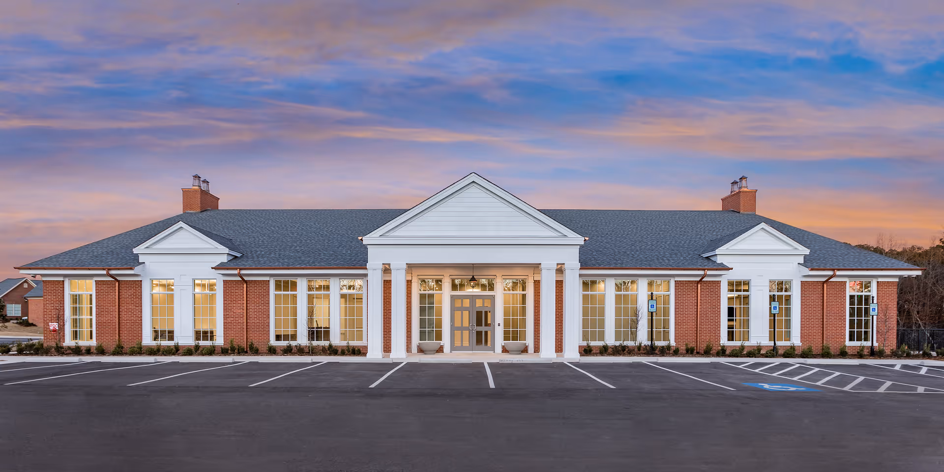 Single-story brick building with white columns and large windows, an empty parking lot in front under a colorful evening sky.