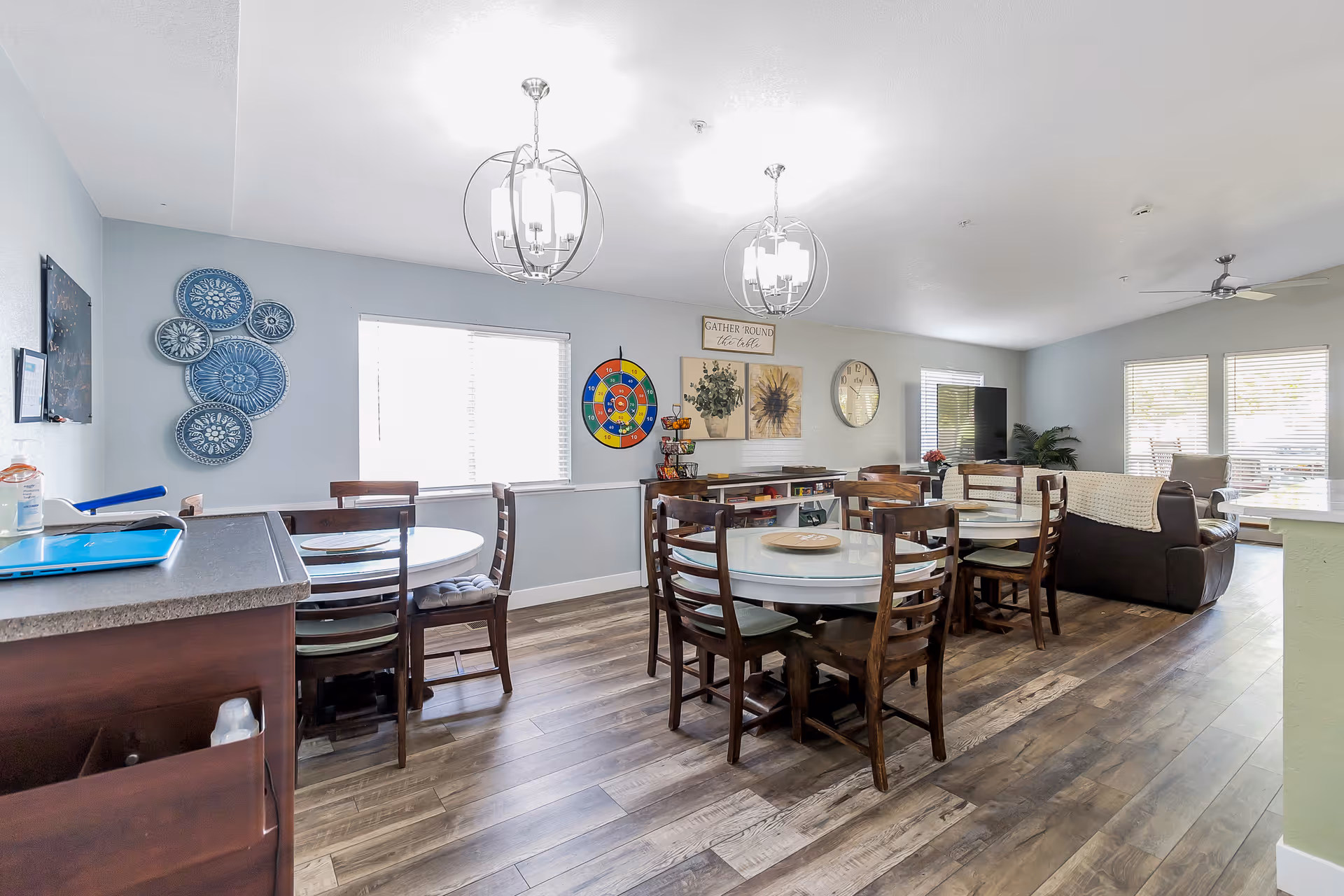 A bright and spacious common area in a senior living facility featuring multiple round tables with wooden chairs, two modern chandeliers hanging from the ceiling, wall decorations including a colorful dartboard and framed artwork, a large clock, and a seating area with a brown leather couch and a TV near large windows with blinds.