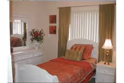 A small bedroom with a single bed covered in red and orange bedding, a green decorative pillow, a white nightstand with a lamp, a white dresser with a vase of flowers, and two framed pictures on the wall. The window has vertical blinds and green curtains.