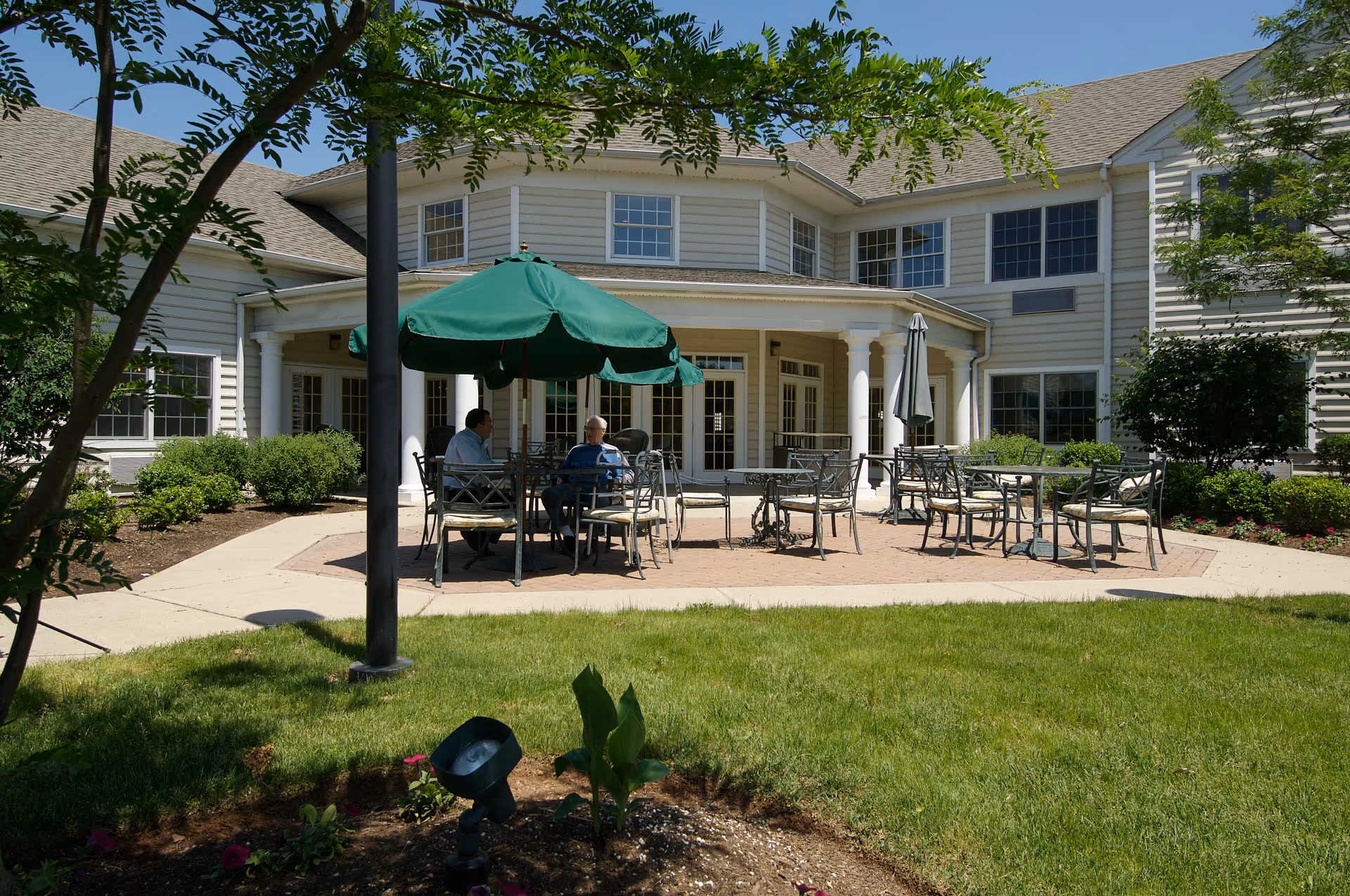 Outdoor patio area at Spring Hills Mount Vernon with metal tables and chairs, some shaded by green umbrellas. Two people are seated and conversing. The patio is surrounded by a well-maintained lawn, bushes, and trees, with a large building featuring multiple windows and white columns in the background.