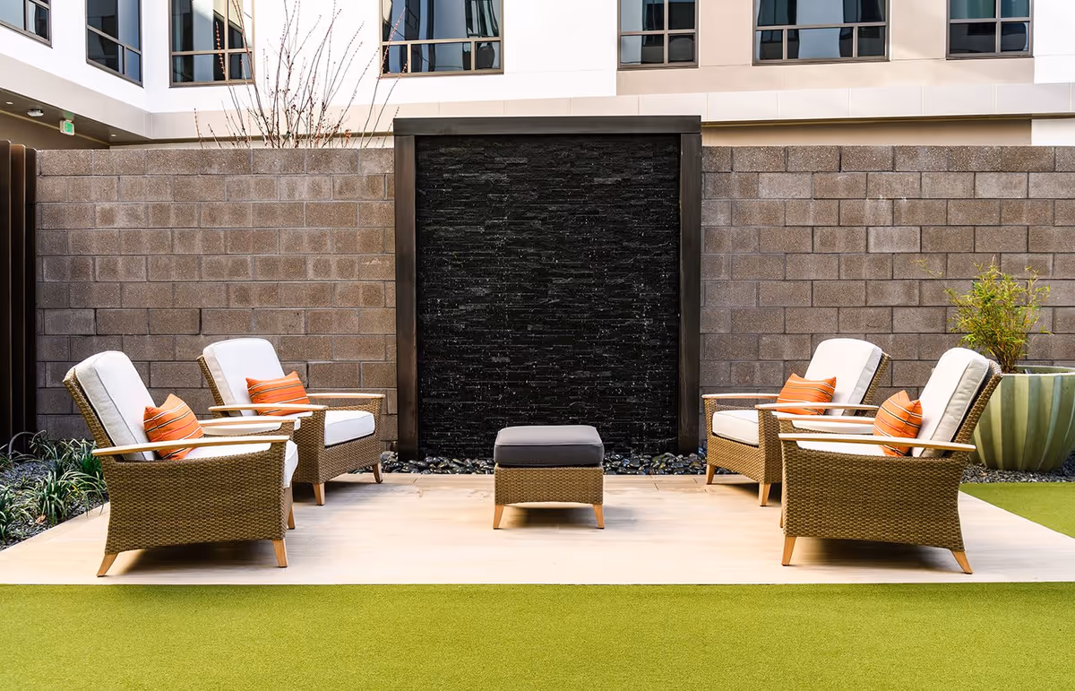 Outdoor courtyard with four wicker lounge chairs and orange pillows arranged around a central ottoman in front of a black stone water feature.