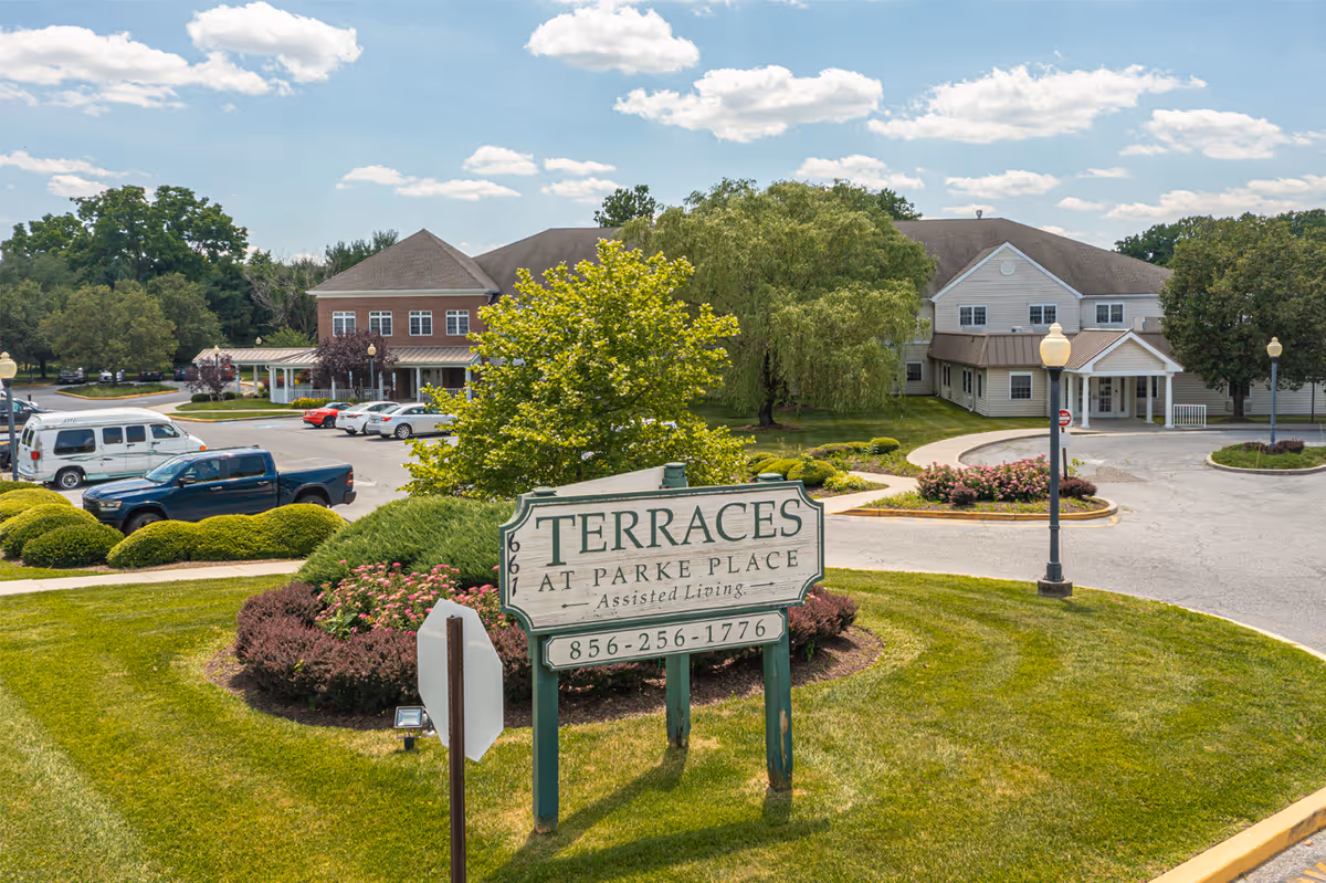 Landscaped entrance with a sign reading "Terraces at Parke Place" in front of an assisted living facility and parking area.