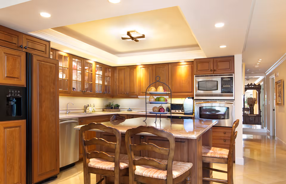 A spacious kitchen with wooden cabinets, a large island with a marble countertop, and three wooden chairs with cushioned seats. The kitchen features built-in stainless steel appliances including a microwave and oven, a refrigerator with a water dispenser, and a dishwasher. The ceiling has recessed lighting and a modern light fixture. There is a three-tiered fruit stand on the island, and the kitchen opens into a hallway with decorative furniture and artwork on the walls.