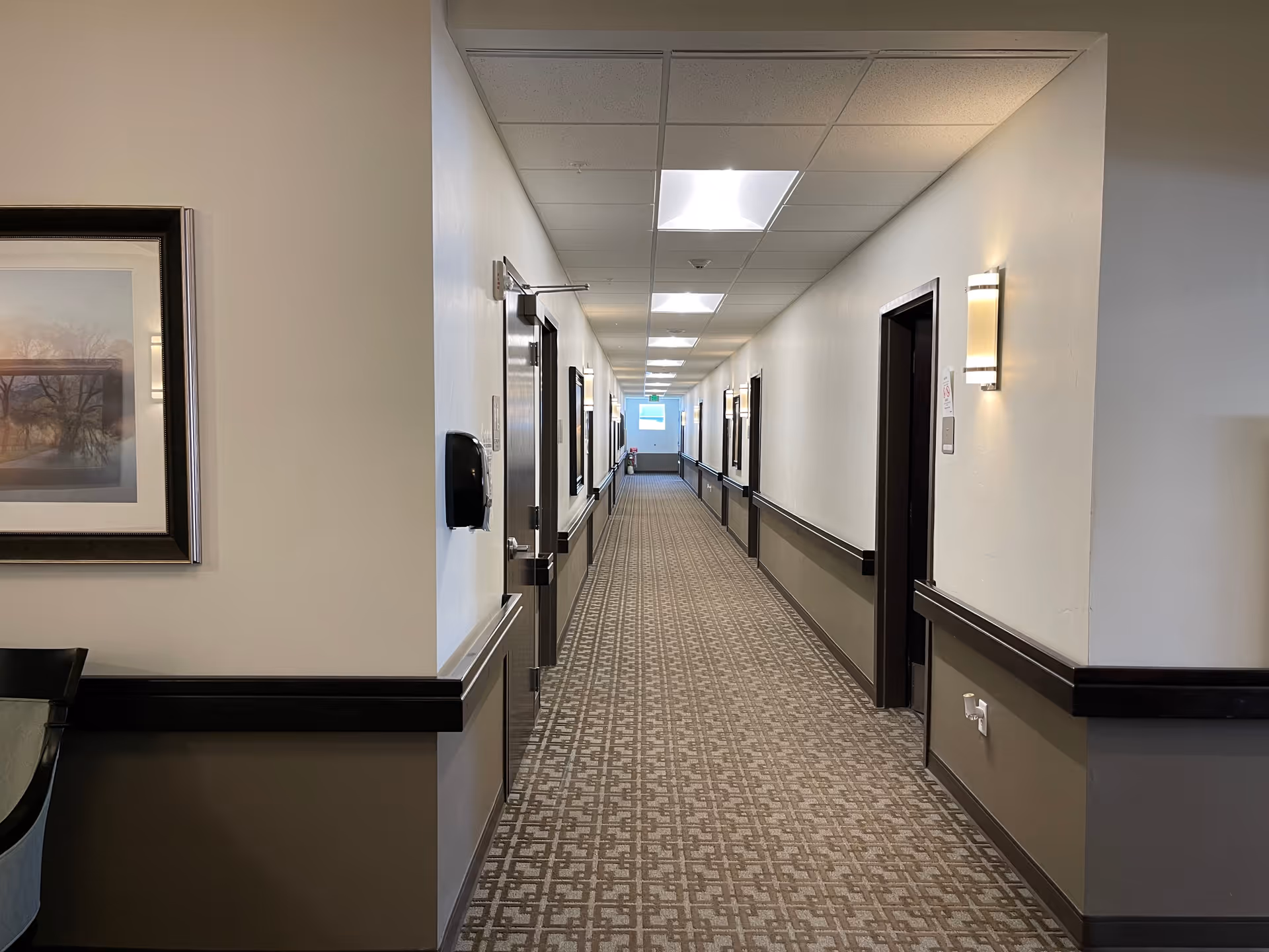A long, well-lit hallway in an assisted living facility with patterned carpet, beige walls with dark brown trim, several doorways on both sides, wall-mounted lights, and a framed picture on the left wall near the foreground.