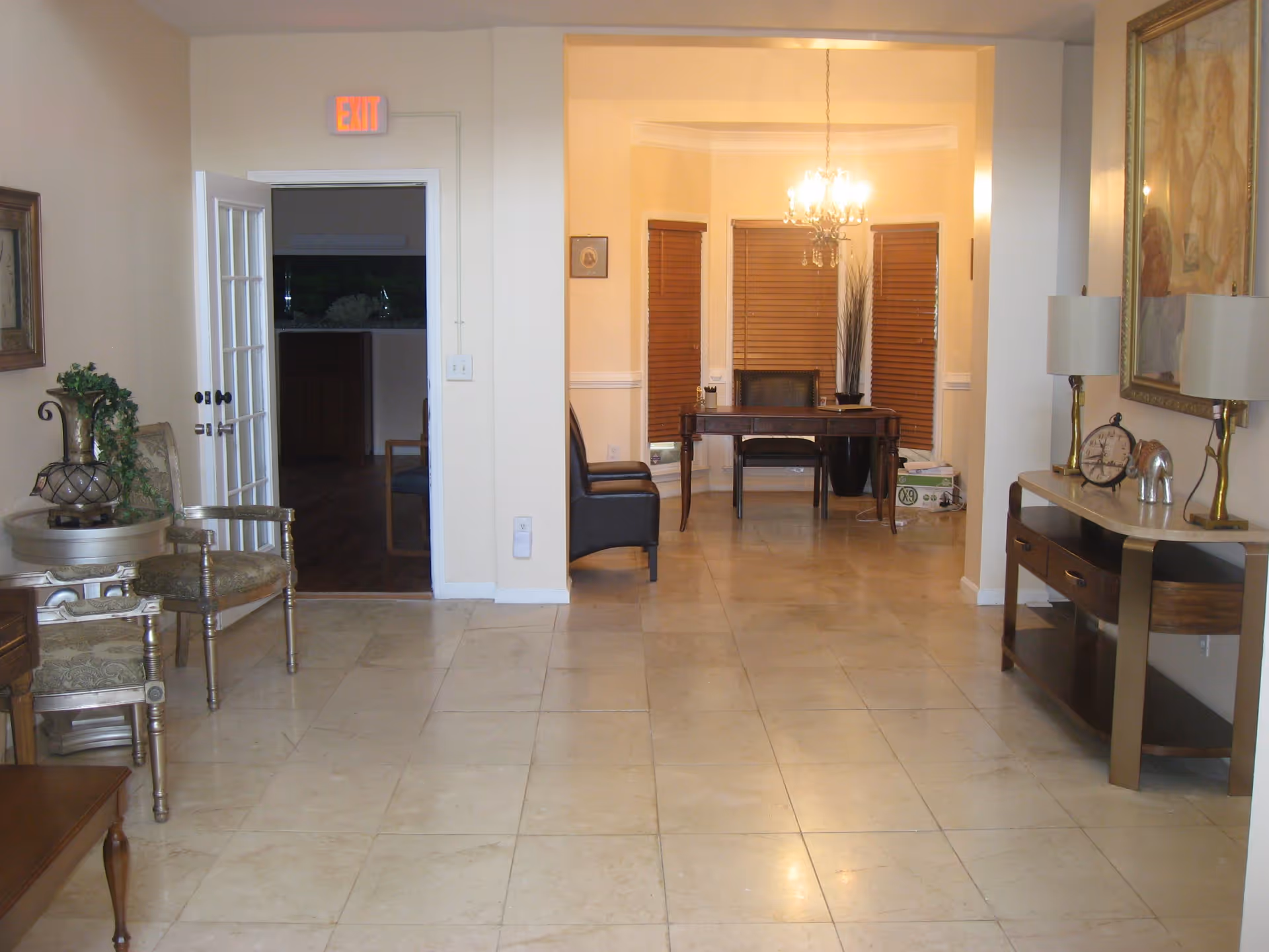 Interior view of a room with beige tiled floor and cream-colored walls. On the left, there are two ornate chairs and a small round table with a decorative plant. In the center background, there is a wooden desk with a chair in front of three windows with closed wooden blinds and a chandelier hanging above. To the right, there is a console table with two lamps, a clock, and a decorative elephant figurine. An exit sign is visible above an open door on the left side.