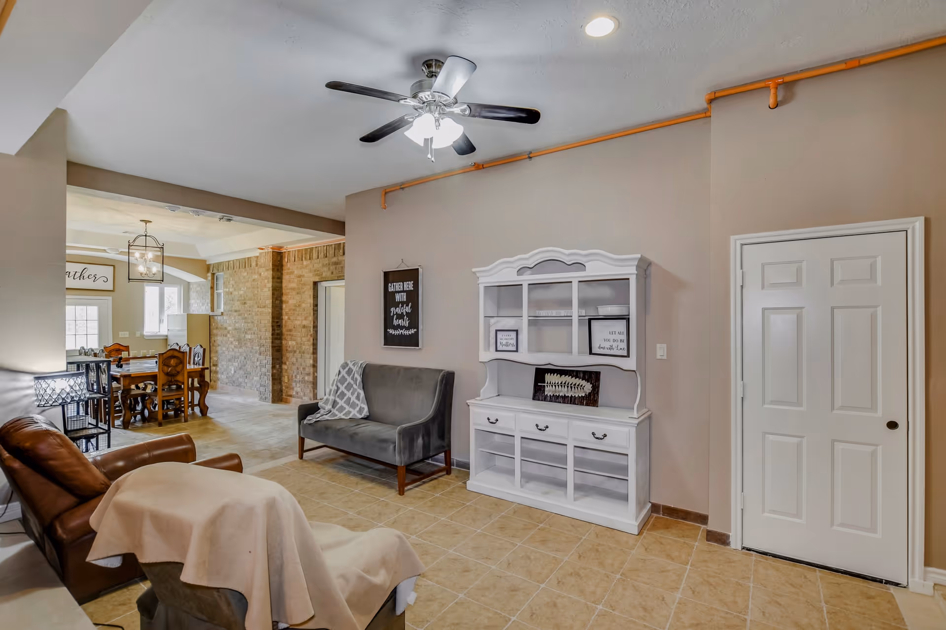 A cozy living room area with a ceiling fan, a gray loveseat, a brown leather recliner covered with a beige blanket, and a white cabinet with shelves and drawers. The room has beige tiled flooring and light brown walls with an orange pipe running along the ceiling. In the background, there is a dining area with a wooden table and chairs, a brick accent wall, and large windows letting in natural light.