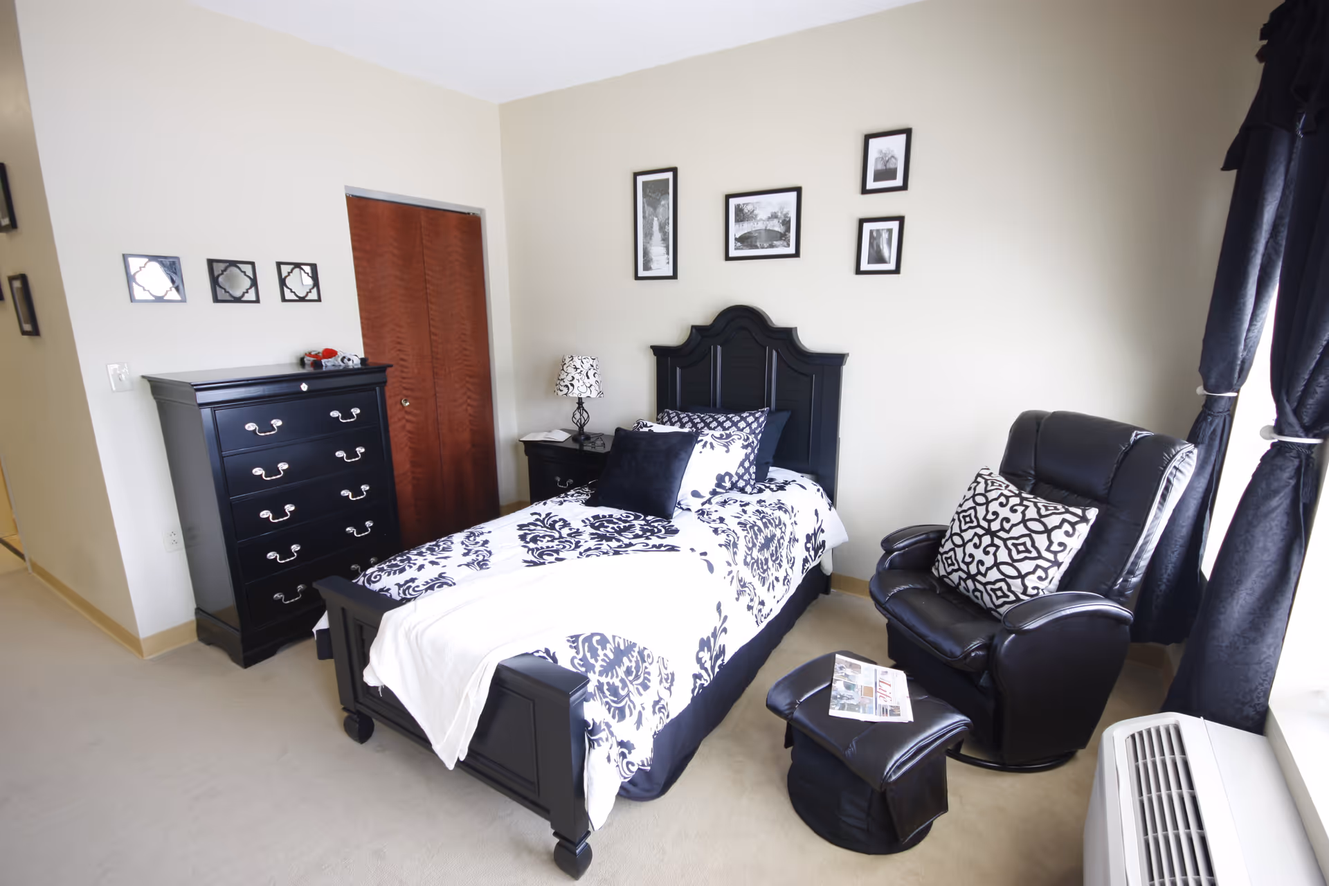 Bright bedroom with a single bed dressed in black-and-white bedding, a black dresser, framed wall art, and a black leather recliner by the window.