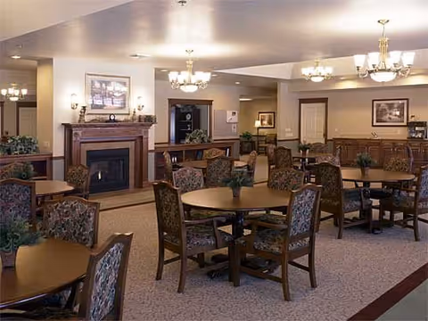 A spacious common area with multiple round wooden tables and upholstered chairs arranged neatly. The room features a fireplace with a framed picture above it, soft lighting from chandeliers, and neutral-colored walls and carpet. There are plants on the tables and decorative elements around the room, creating a warm and inviting atmosphere.