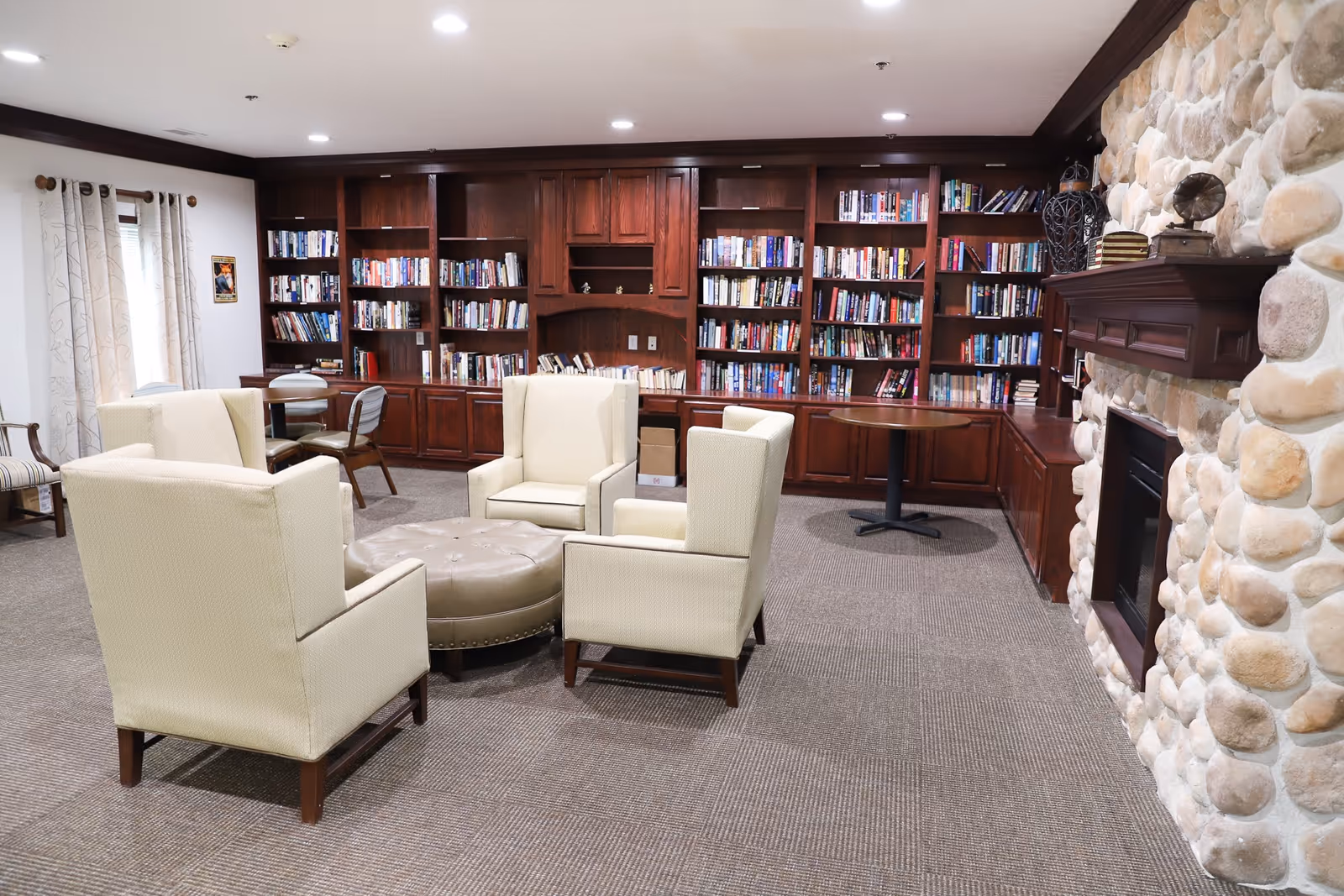 A cozy library room with four beige armchairs arranged around a round, tufted ottoman. The room features a large wooden bookshelf filled with books along the back wall, a stone fireplace on the right, and a small round table with chairs near the window on the left. The carpeted floor and soft lighting create a warm and inviting atmosphere.