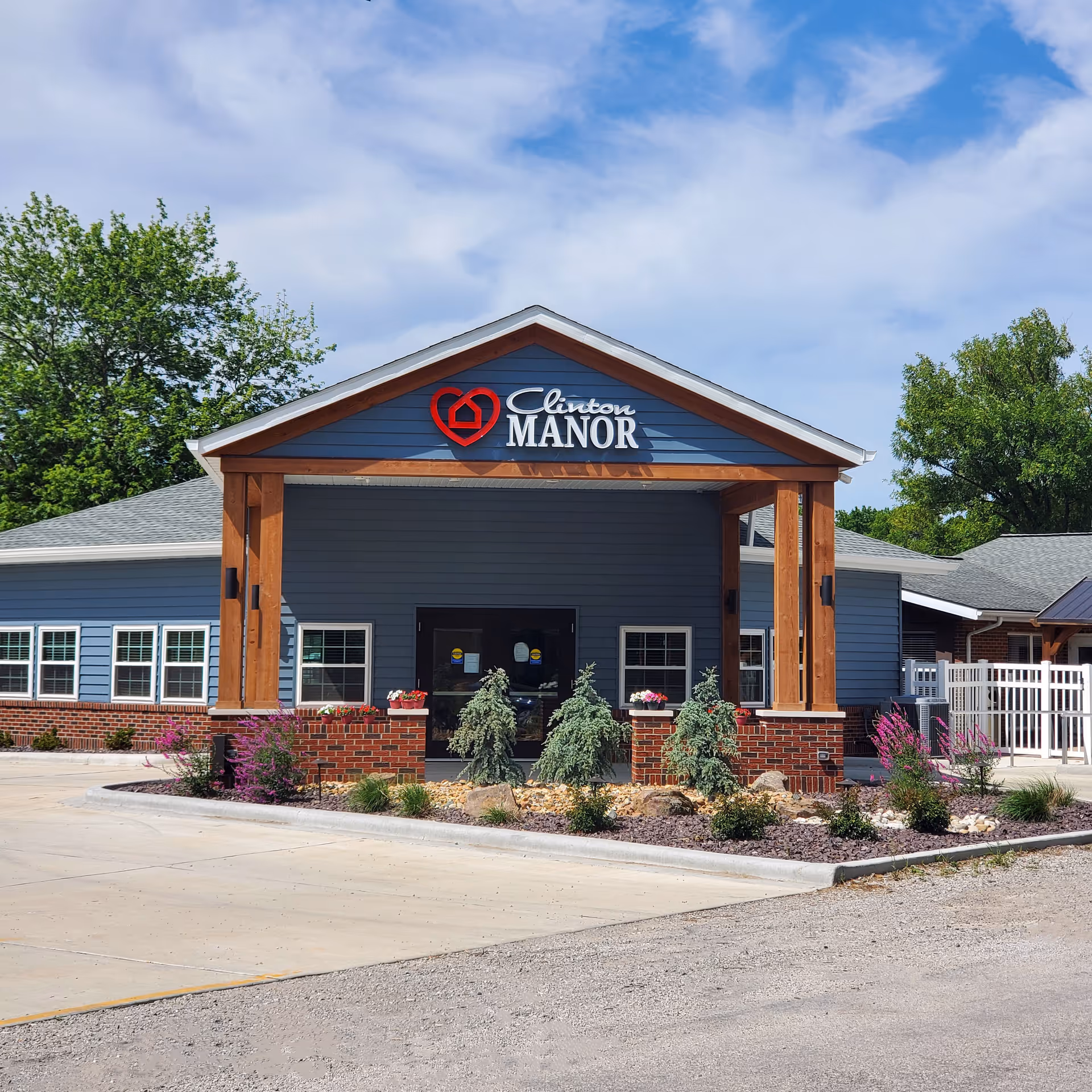 Front exterior view of Clinton Manor Living Center building with a covered entrance supported by wooden beams, landscaped with small trees and flowers, under a partly cloudy blue sky.