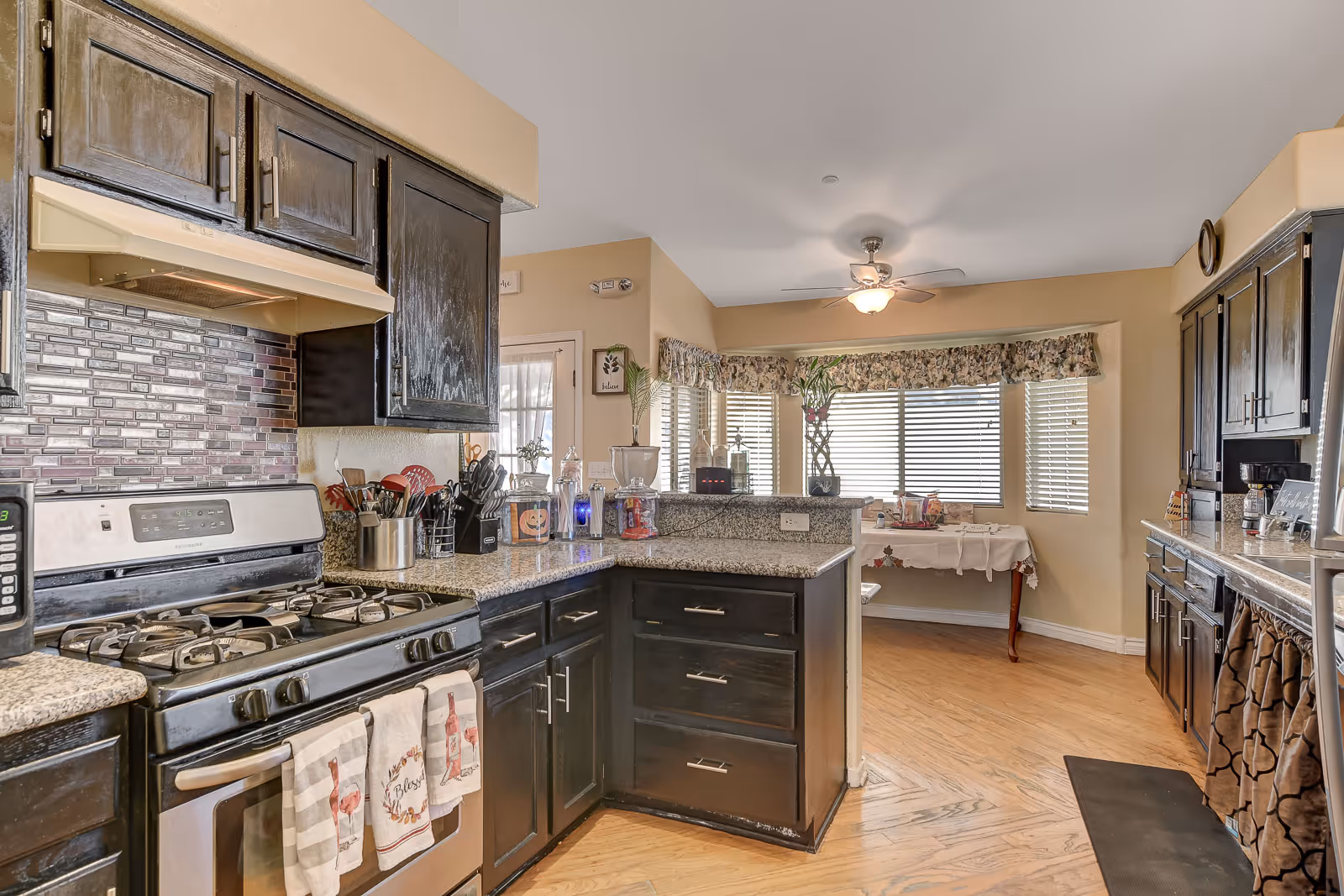 A bright kitchen with dark wooden cabinets, a stainless steel gas stove with three decorative towels hanging on the handle, granite countertops, and a mosaic tile backsplash. The kitchen opens into a dining area with large windows covered by blinds and floral valances, a ceiling fan with light, and a table with a white tablecloth near the windows.