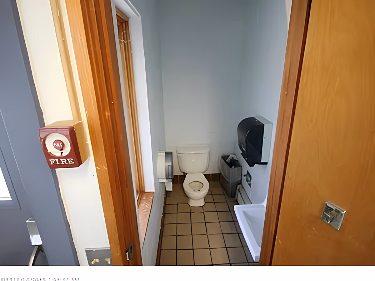 Small bathroom with a white toilet, a white sink, a black paper towel dispenser, a trash bin, and a window with wooden trim. The floor is tiled with brown square tiles, and there is a fire alarm pull station mounted on the wall outside the bathroom.