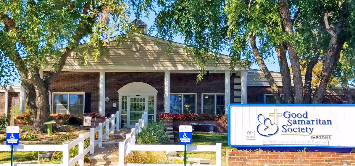 Exterior view of Good Samaritan Society - Parsons building with a brick facade, white columns, and a glass entrance door. There are large trees and landscaped greenery around the entrance, along with a white fence and signs indicating handicapped parking. A large sign with the facility's name is visible on the right side of the image.