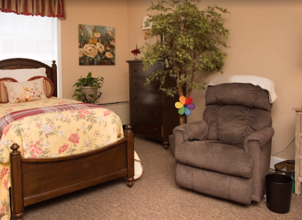 A cozy bedroom with a wooden bed frame featuring floral bedding and decorative pillows. Next to the bed is a potted plant on a small stand. A dark wooden chest of drawers stands against the wall with a lamp and small flower vase on top. A large artificial tree with a colorful pinwheel is beside a plush brown recliner chair with a white throw draped over the back. The room has beige walls and carpeted floor.
