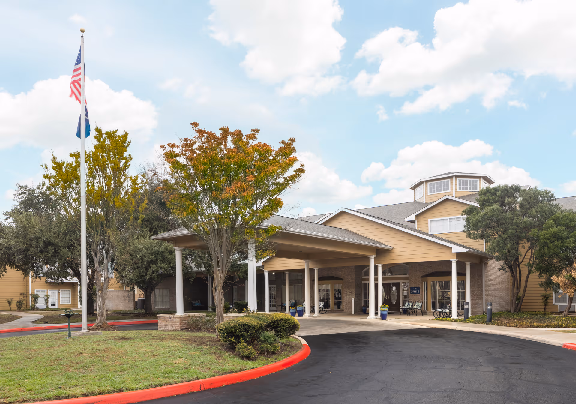 Front entrance of a senior living building with a covered drop-off area, flagpole, and landscaped driveway.