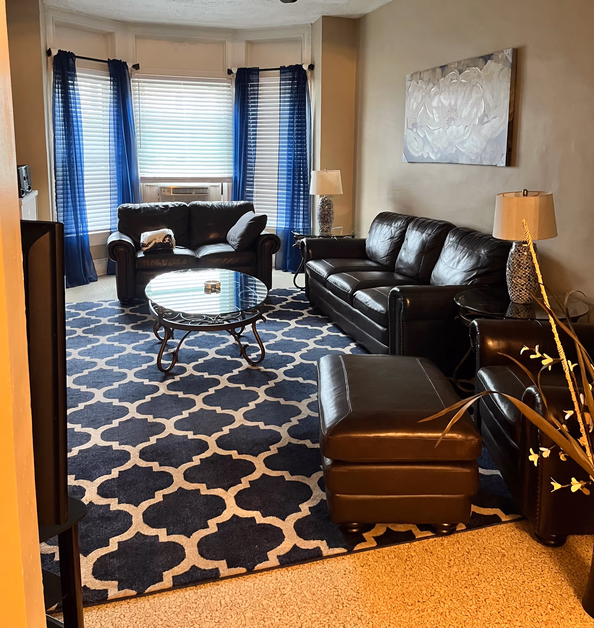A furnished living room with black leather sofas and ottoman, a glass coffee table on a blue patterned rug, lamps, wall art, and bay windows with blue curtains.