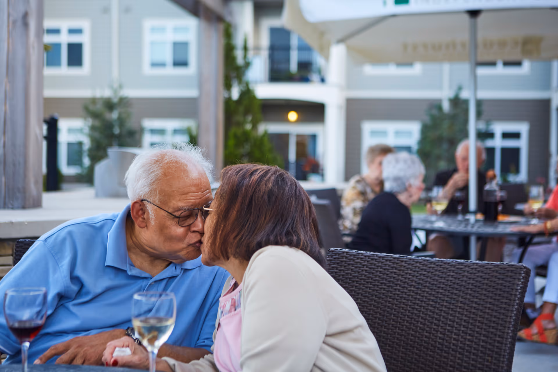 An elderly couple sharing a kiss while sitting at an outdoor dining area with wine glasses on the table. In the background, other elderly people are seated at tables under large umbrellas, with a residential building visible behind them.