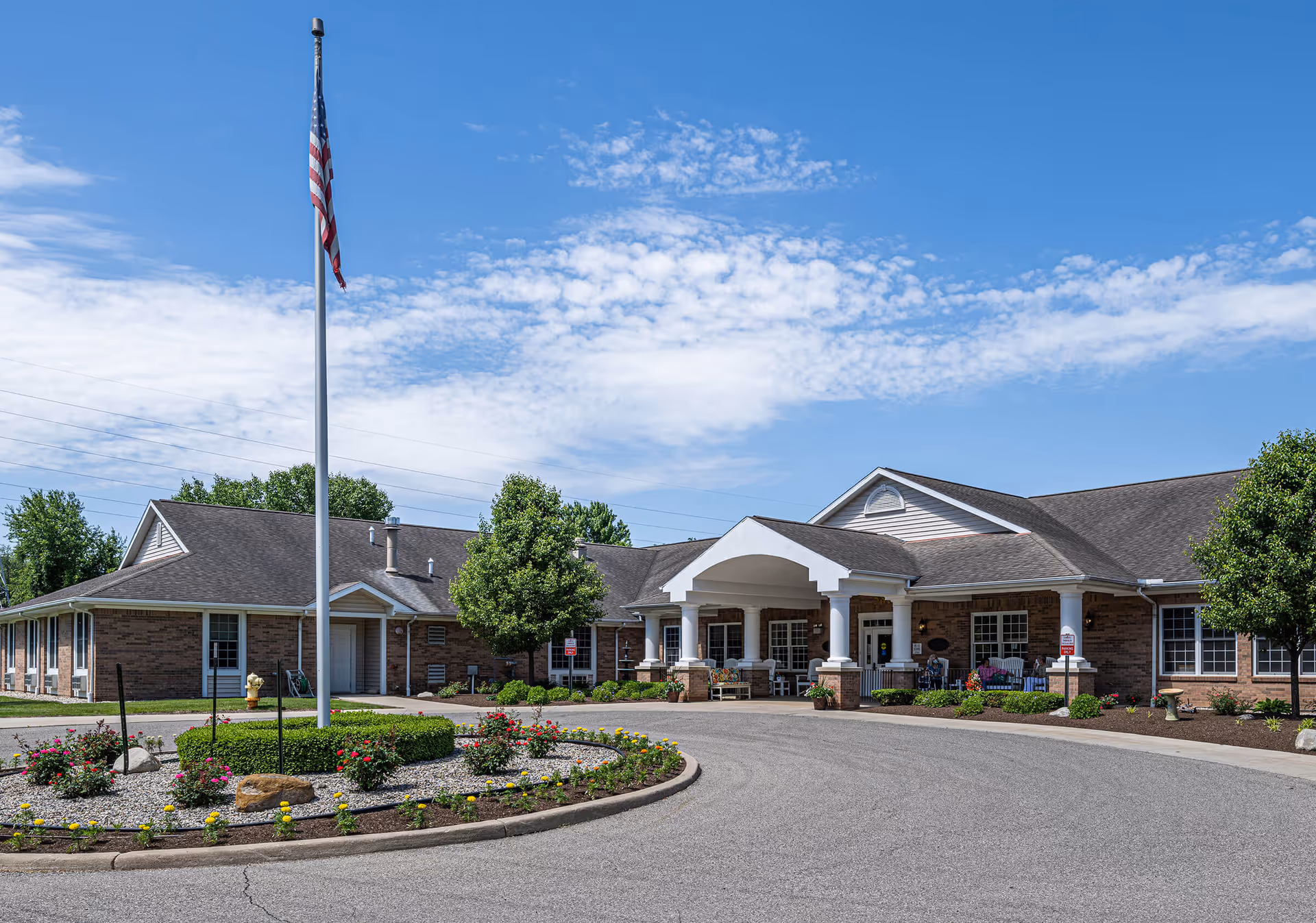 Exterior view of a single-story senior living community building with a covered entrance, surrounded by landscaped flower beds and trees, under a partly cloudy blue sky. An American flag is flying on a flagpole in front of the building.