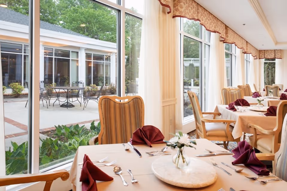 A bright dining room with large windows overlooking an outdoor patio. Tables are set with beige tablecloths, burgundy folded napkins, silverware, and small flower vases. The chairs have wooden frames with striped upholstery, and the room features patterned curtains and a carpeted floor.