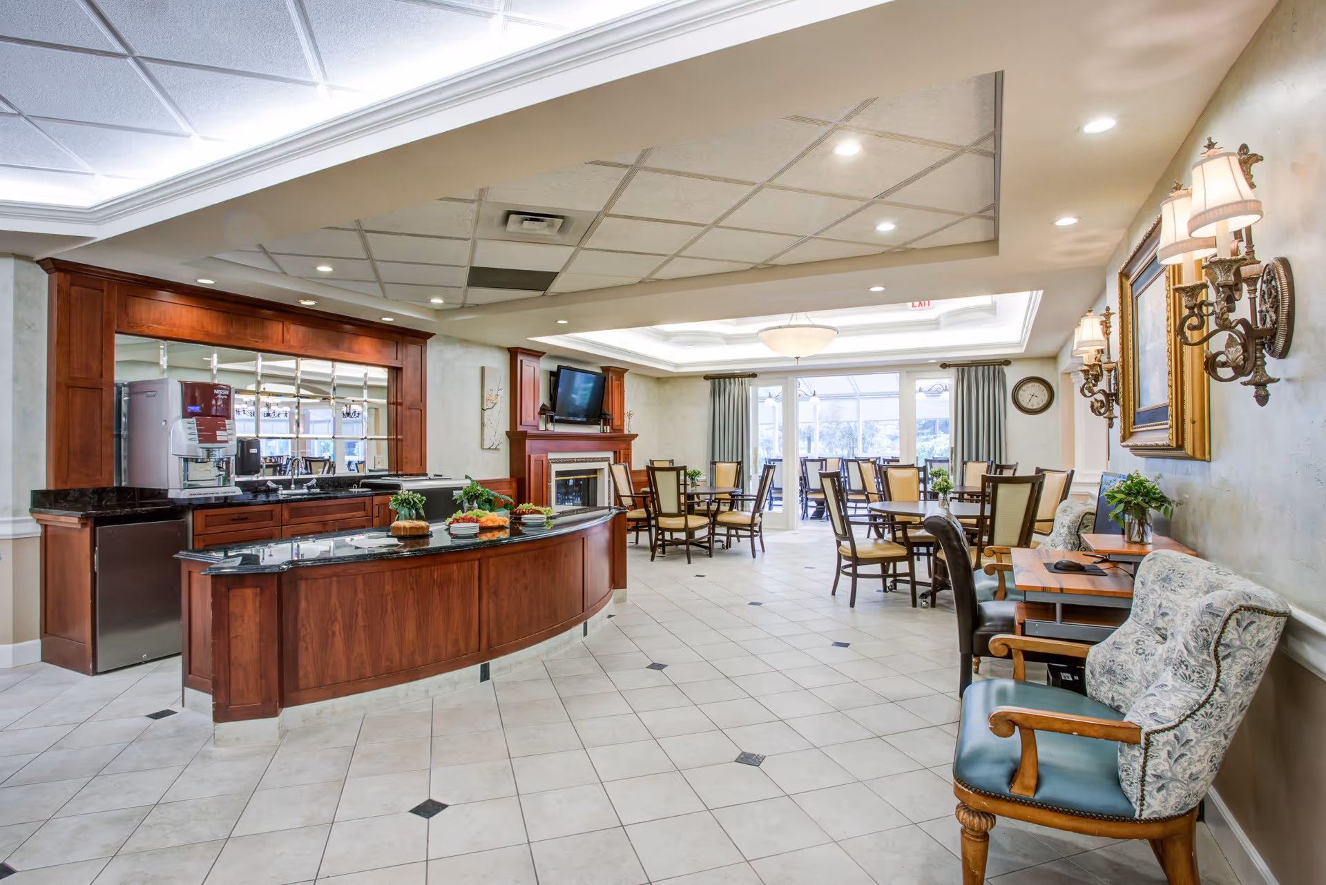 A spacious dining area in The Village Senior Residence featuring multiple wooden tables and chairs arranged neatly. There is a curved wooden counter with a black countertop displaying plates and food items. The room has a tiled floor, a ceiling with recessed lighting, and a wall-mounted TV above a fireplace. On the right side, there are upholstered chairs and a small desk with a computer. Large windows with curtains allow natural light to fill the space.