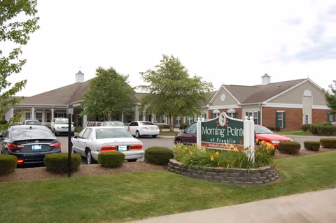 Exterior view of Morning Pointe of Franklin senior living facility showing a single-story brick and siding building with a parking lot in front. Several cars are parked, and there is a landscaped area with bushes and a sign that reads 'Morning Pointe of Franklin'. Trees and a partly cloudy sky are visible in the background.