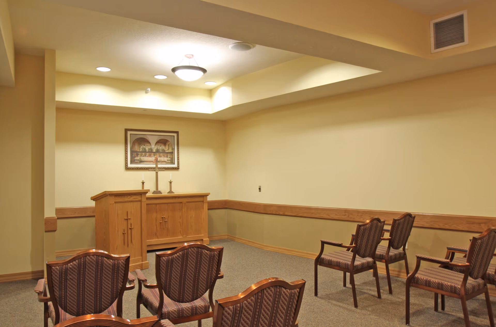 A small chapel or prayer room with wooden chairs arranged in rows facing a wooden altar with a cross and two candles. A framed religious painting hangs on the wall behind the altar. The room has beige walls and ceiling with recessed lighting.
