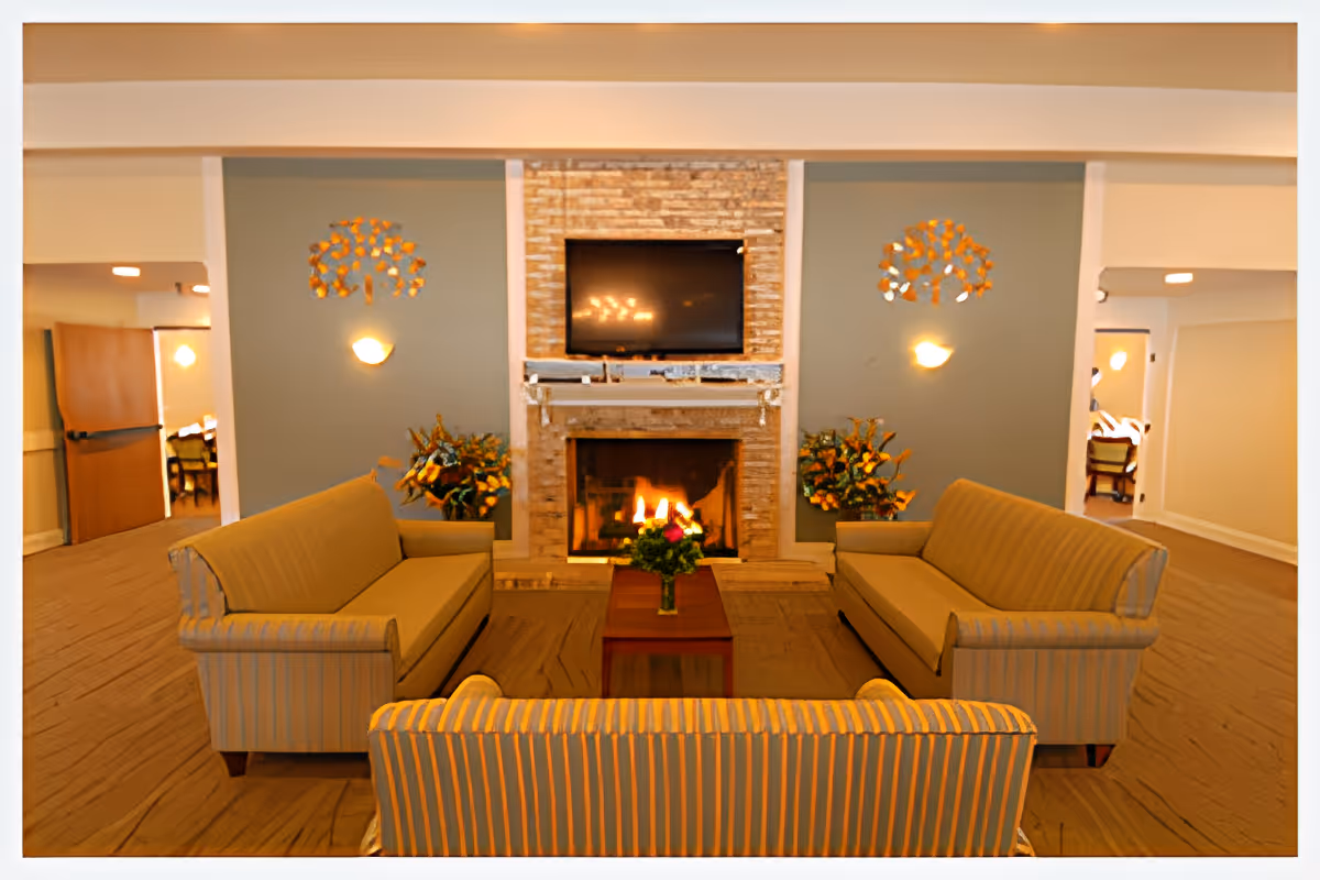 A cozy living room area in Delta Retirement Center featuring three striped sofas arranged around a wooden coffee table with a flower vase. A lit fireplace with a stone surround is centered on the far wall, above which a flat-screen TV is mounted. Two wall-mounted light fixtures and decorative wall art flank the fireplace. The room has beige carpeting and soft lighting, with doorways visible on either side leading to other rooms.