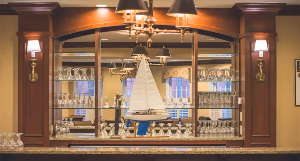 A polished wooden bar area with glass shelves holding various glassware including wine glasses and tumblers. A decorative model sailboat is displayed in the center. The background shows windows with white shutters and wall sconces with lampshades on either side of the bar.