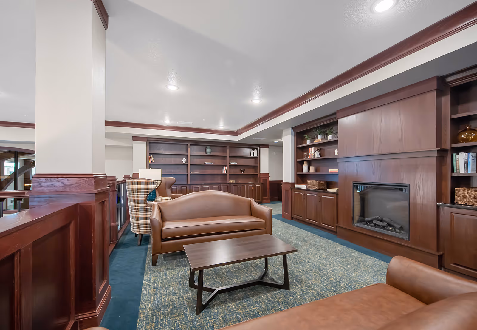 A cozy senior living common area with brown leather sofas, a wooden coffee table, built-in wooden bookshelves, and a fireplace. The room features a blue patterned carpet and recessed ceiling lights.