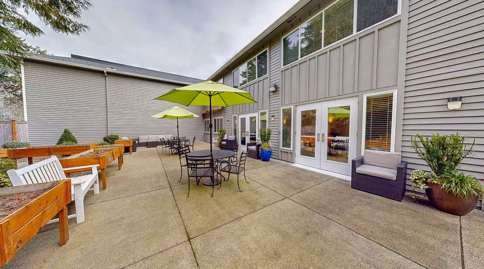 Outdoor courtyard patio at a senior living facility with round tables and green umbrellas, raised planter boxes, and a building facade with glass doors.