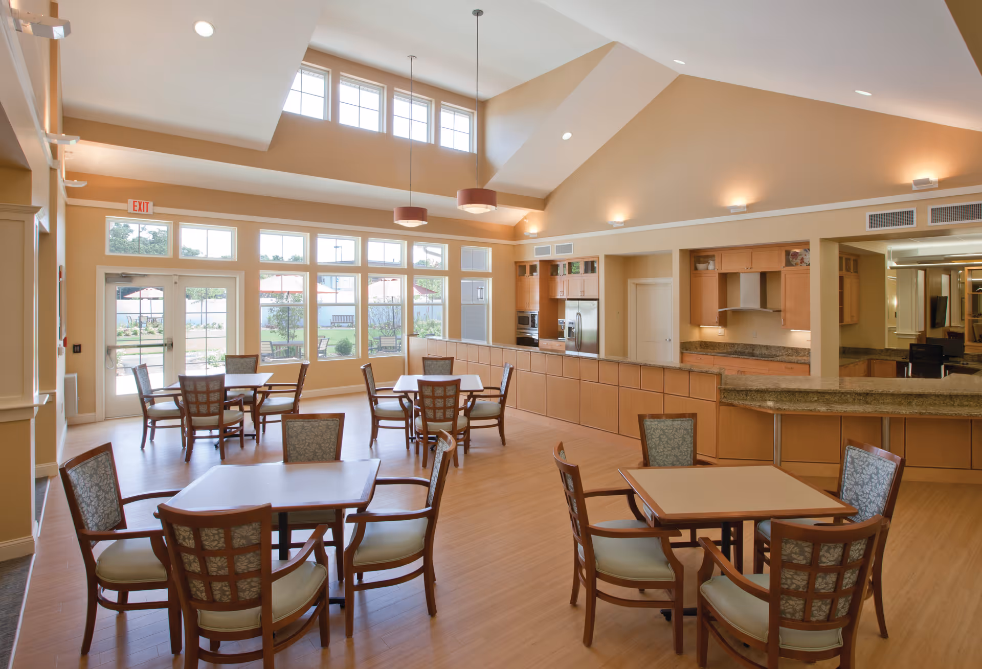 Sunlit communal dining area with wooden tables and chairs, large windows, and an open kitchen under a vaulted ceiling.