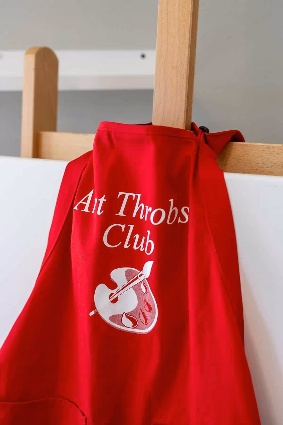A red apron hanging on a wooden easel with the words 'Art Throbs Club' and an image of a painter's palette and brush printed on it.