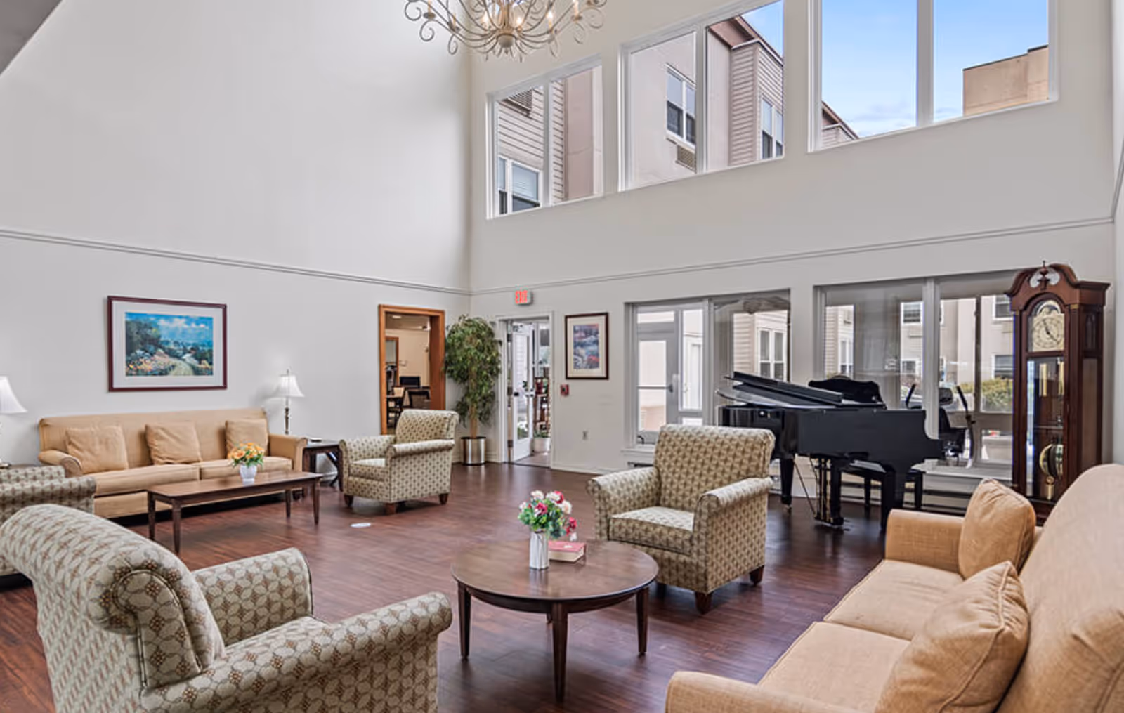 A spacious living room with high ceilings and large windows letting in natural light. The room is furnished with beige sofas, patterned armchairs, a round wooden coffee table with flowers and books, a grand piano, a tall grandfather clock, and framed artwork on the walls. The floor is wooden, and there is a chandelier hanging from the ceiling.