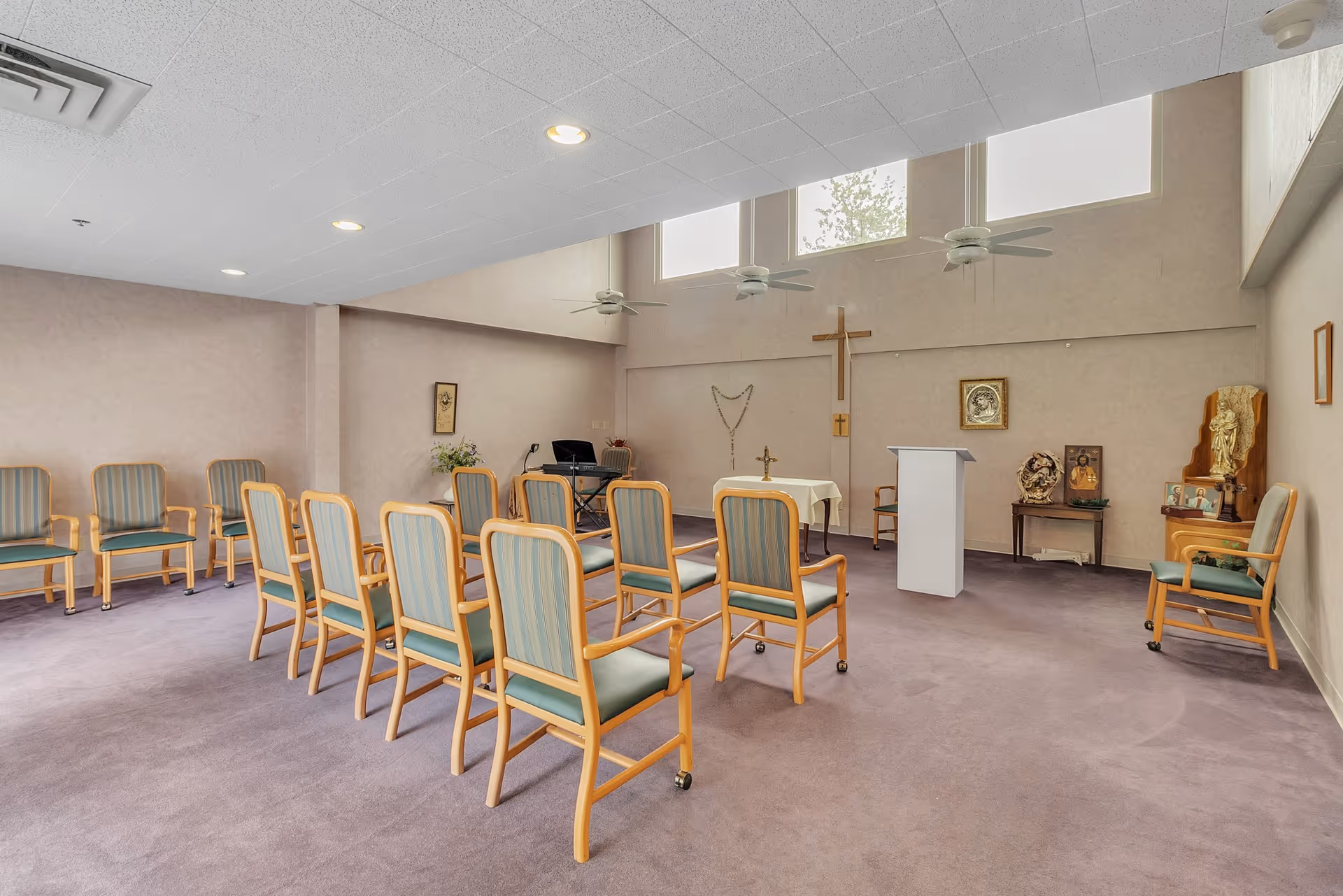 A small chapel or prayer room with rows of wooden chairs with green cushions arranged facing a white podium and altar. The room has a purple carpet, beige walls, and three ceiling fans. Religious items including a large wooden cross, a statue, framed pictures, and a rosary are displayed on the walls and tables. Natural light enters through high windows near the ceiling.