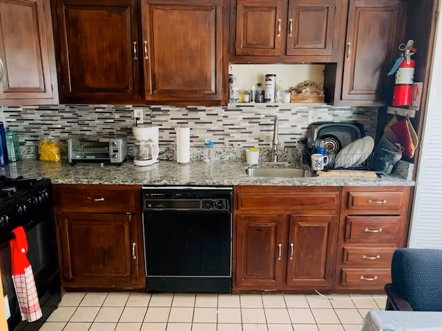 Kitchen with dark wooden cabinets, a granite countertop, a tiled backsplash in shades of gray and white, a black dishwasher, a stainless steel sink with a faucet, a coffee maker, a toaster oven, paper towels, and a drying rack with dishes. A fire extinguisher is mounted on the wall to the right.