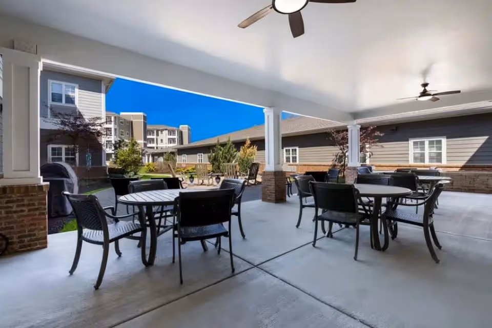 Covered outdoor patio area with several round tables and black chairs arranged for seating. Ceiling fans are mounted on the white ceiling. The patio overlooks a courtyard with greenery and buildings in the background under a clear blue sky.