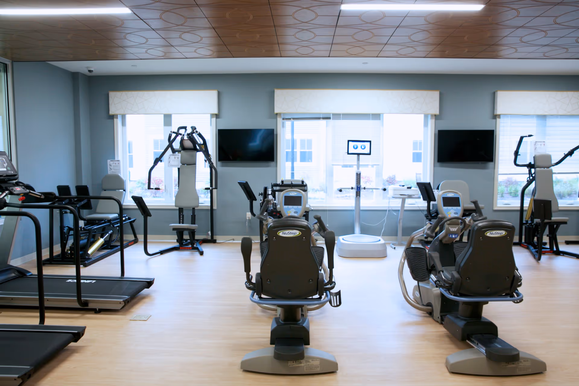 Bright fitness room with treadmills, recumbent bikes and strength equipment arranged facing windows and TVs.