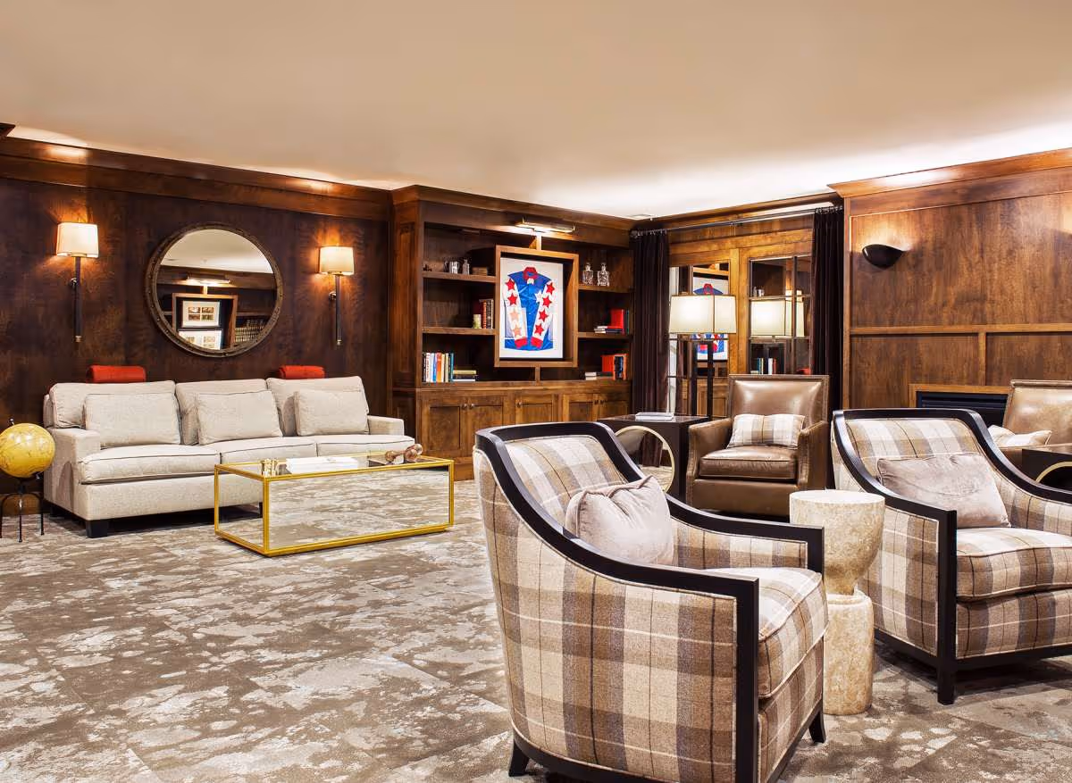 A cozy living room with a beige sofa, two plaid armchairs, and two brown leather chairs arranged around a mirrored coffee table. The room features dark wood paneling, built-in shelves with books and decor, a round mirror on the wall, and warm lighting from wall sconces and a floor lamp.