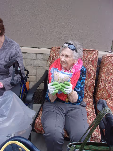 An elderly woman wearing a bright pink scarf, green gloves, and sunglasses on her head is sitting on a patterned outdoor chair, smiling and holding a colorful pillow. Another person is partially visible sitting next to her, and there is a walker and a plastic bag nearby.