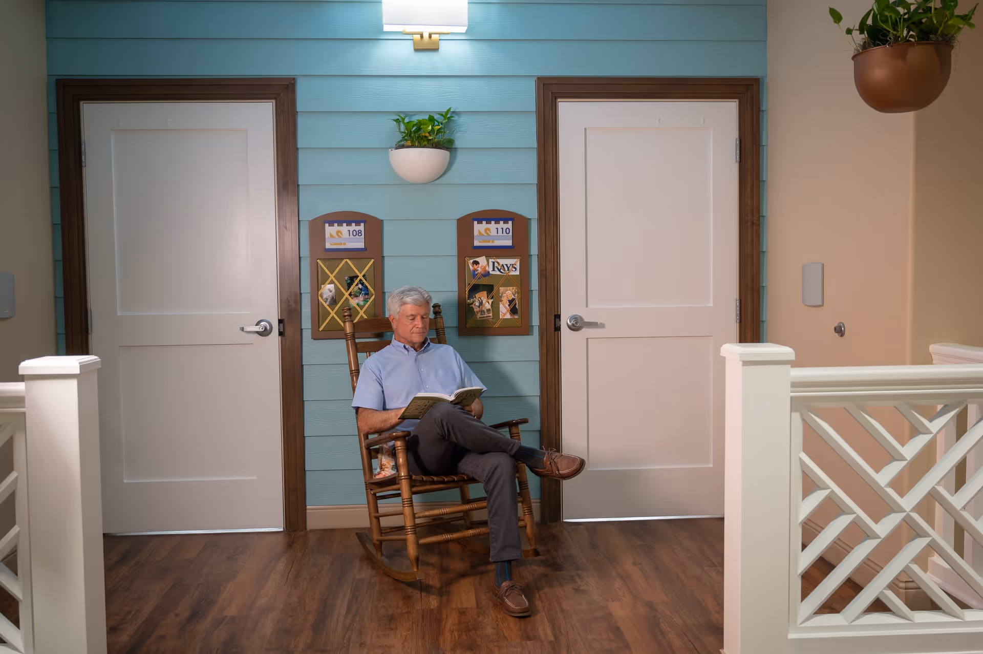 An elderly man with gray hair sits in a wooden rocking chair reading a book in a hallway with two white doors framed in dark wood. The wall behind him is painted light blue with two bulletin boards labeled 108 and 110, decorated with photos and notes. A white wall-mounted planter with green plants is above the bulletin boards, and another hanging planter is visible on the right side. The floor is wood, and white railings with a crisscross pattern are in the foreground.