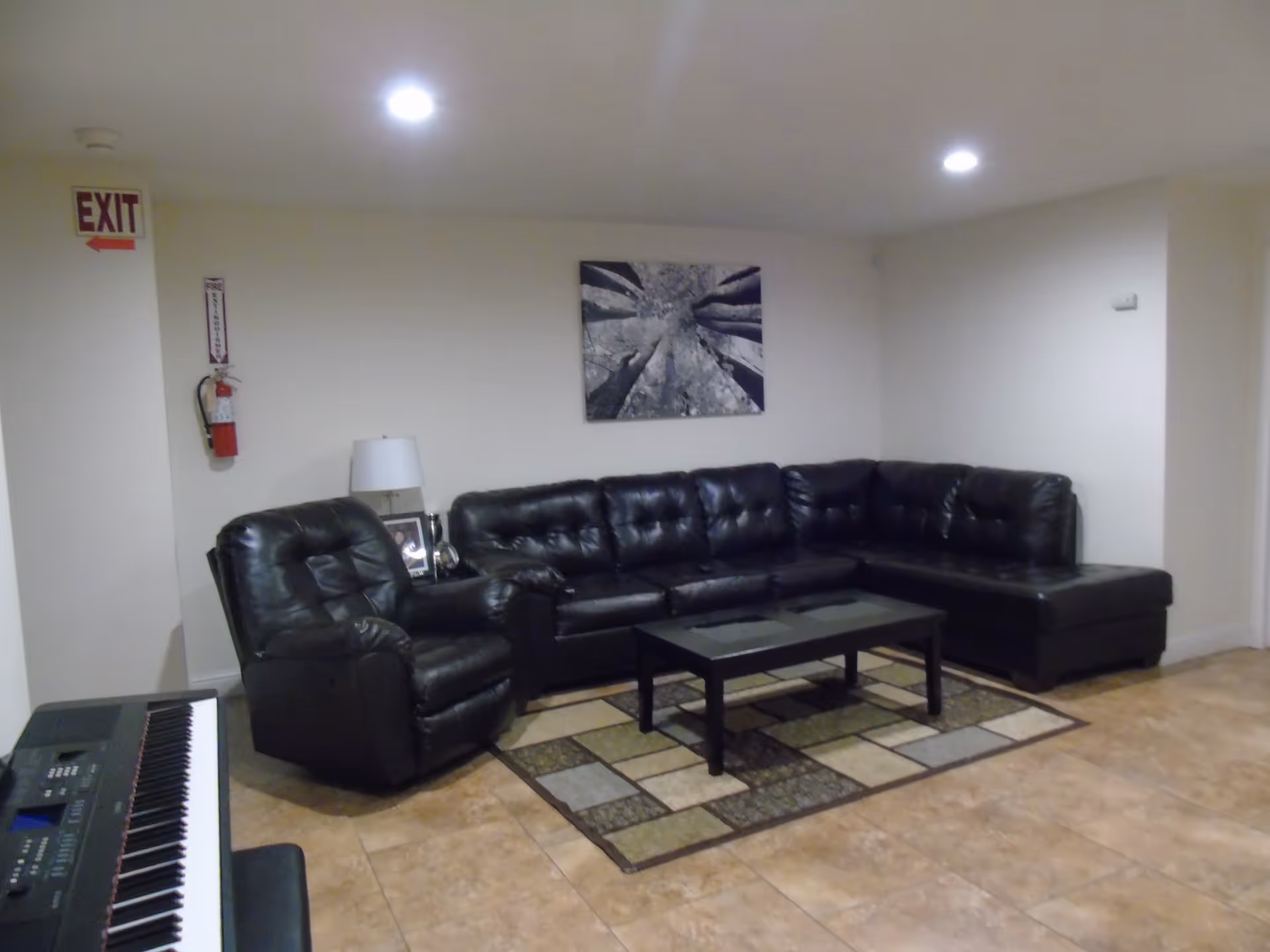 Small sitting area with a black leather sectional sofa and recliner, coffee table on an area rug, and a keyboard against a tiled floor.