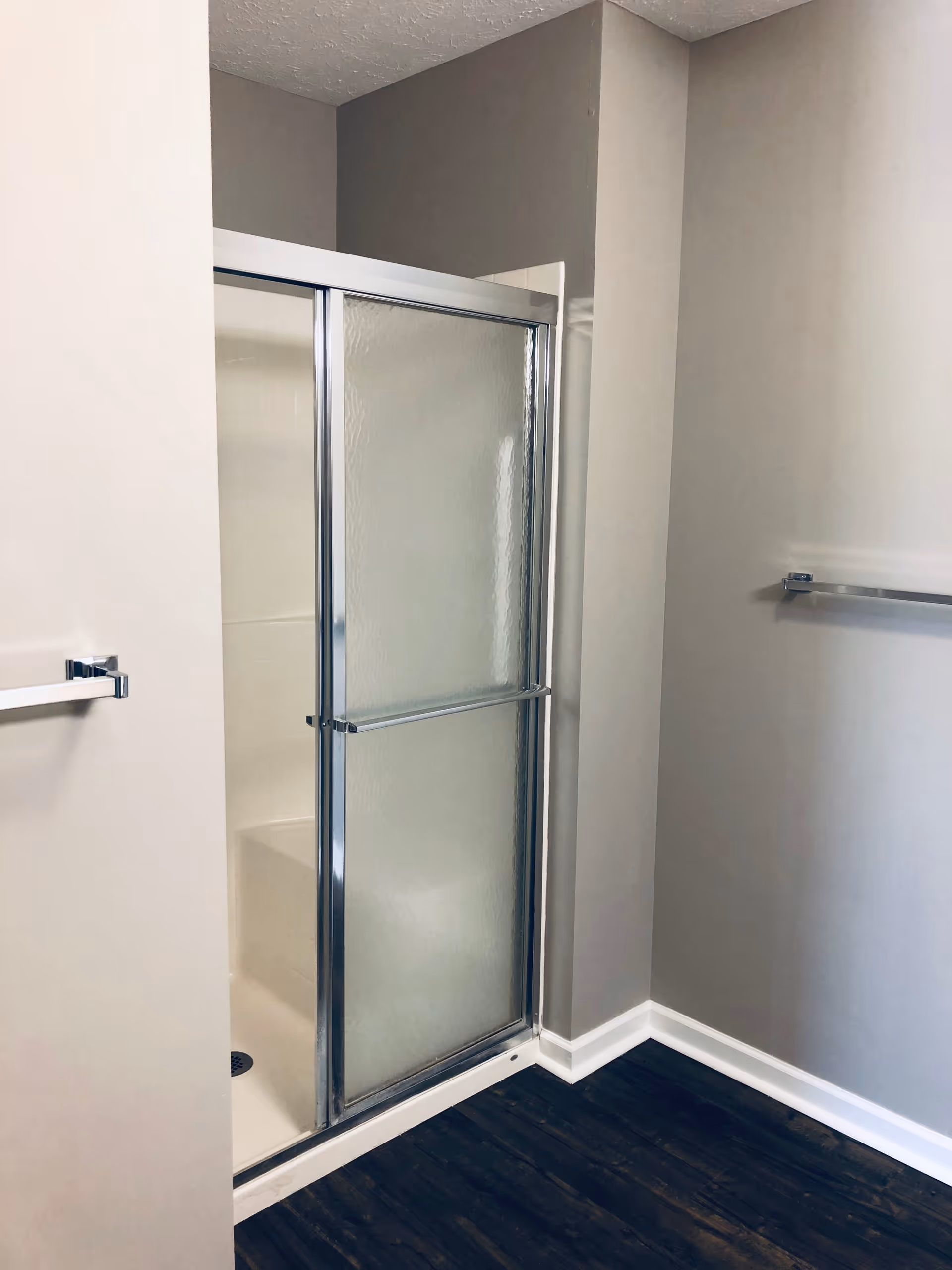 A bathroom corner featuring a shower with frosted sliding glass doors and chrome frame. The walls are painted in neutral tones, and there are two chrome towel bars mounted on the walls. The floor has dark wood-like flooring.