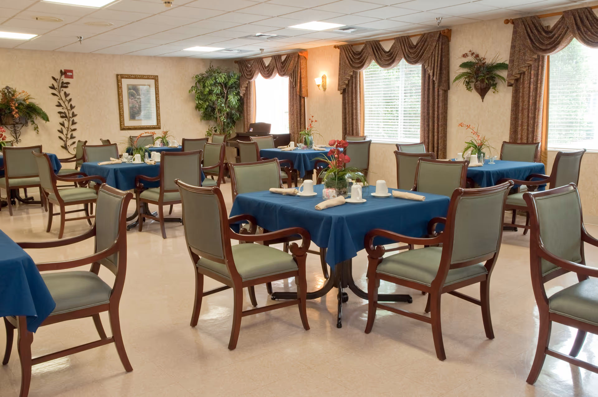 A dining room with multiple tables covered in blue tablecloths, each set with cups, napkins, and small floral centerpieces. The room has green cushioned wooden chairs, large windows with brown curtains, and decorative plants and wall art.