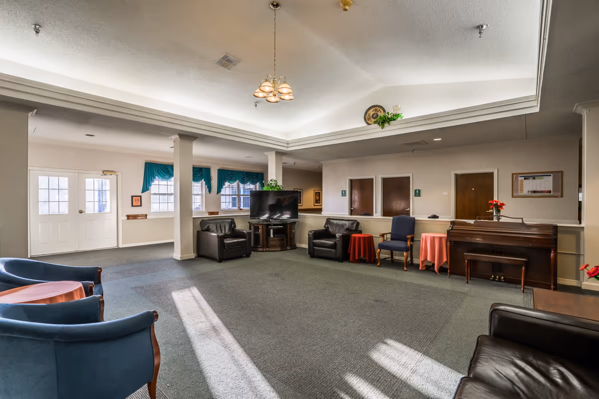 A spacious common area in Saunders House featuring multiple seating options including blue and black armchairs, a wooden piano with a bench, small round tables with red tablecloths, a flat-screen TV on a wooden stand, and windows with teal curtains letting in natural light.