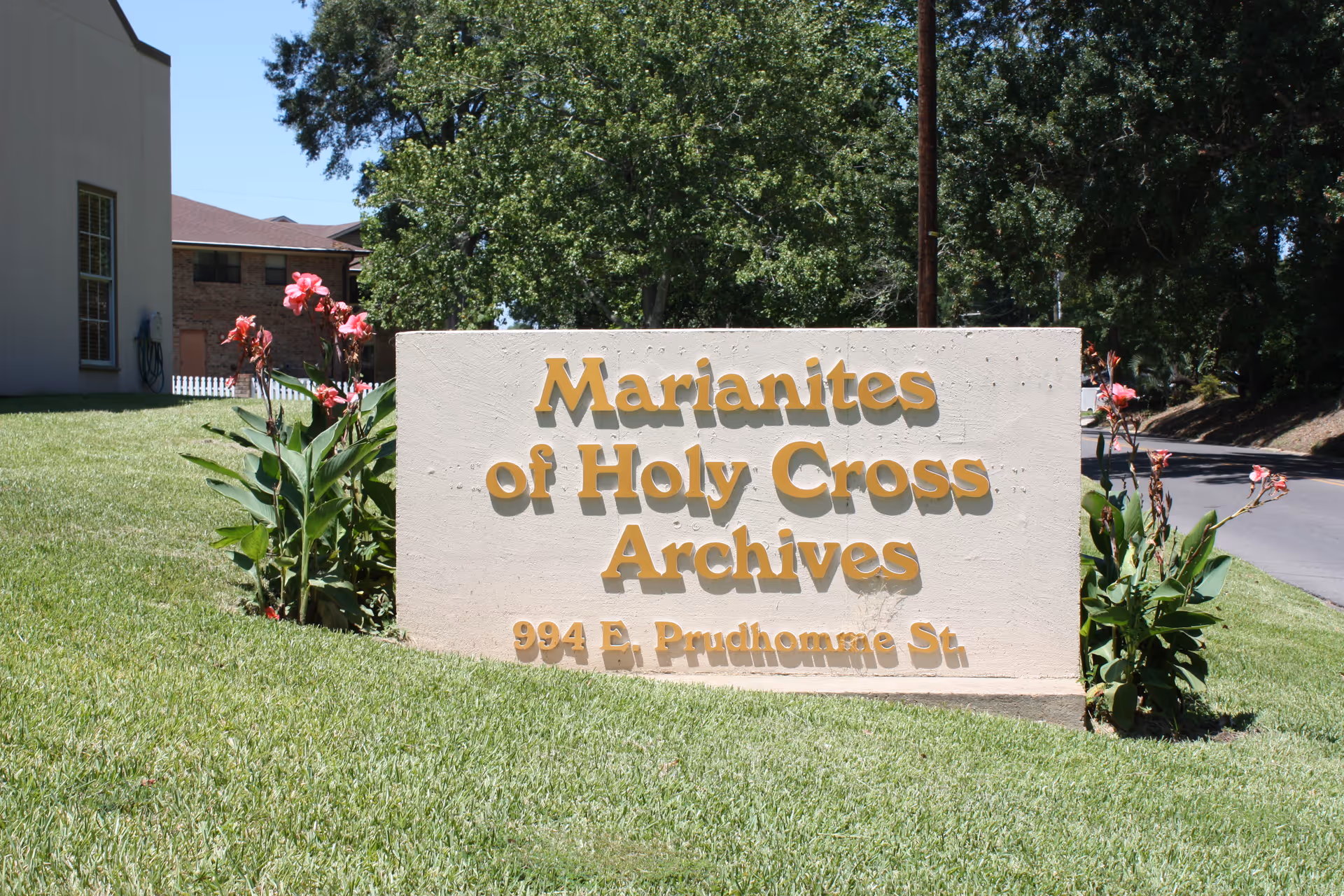 Outdoor image of a beige stone sign with yellow letters that reads 'Marianites of Holy Cross Archives 994 E. Prudhomme St.' The sign is surrounded by green grass and flowering plants, with trees and a road visible in the background.