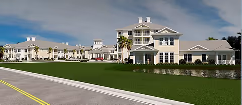 A row of light-colored senior living buildings with a pond and palm trees along a road