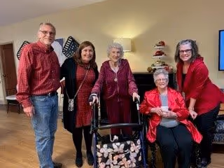 A group of five people posing indoors in a living room setting. Three women and one man are standing, while one woman is seated. The woman in the center uses a walker. The room has warm lighting, a lamp, and decorative items on a shelf in the background.