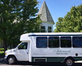 A white and green Alderwood Assisted Living shuttle bus parked on a street with a building featuring a pointed turret and surrounded by green trees in the background under a clear blue sky.